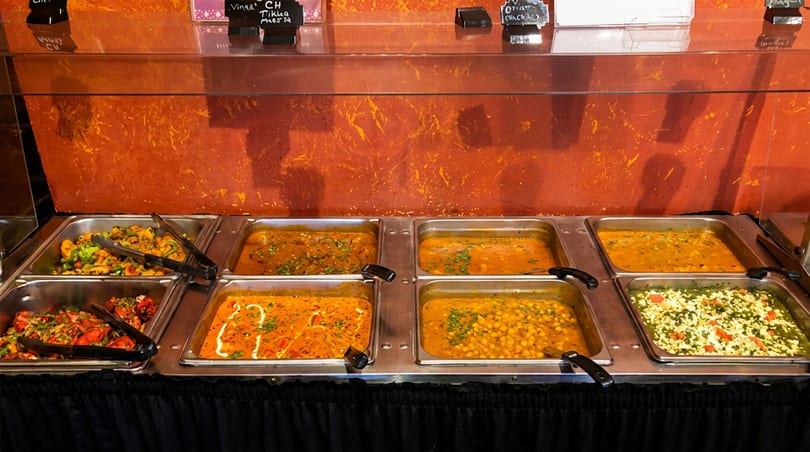 Indian buffet line with multiple hot trays of colorful curries, vegetables, and paneer dishes.